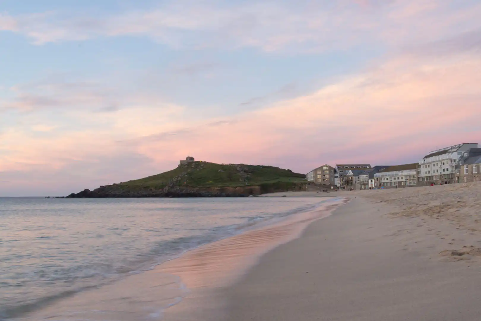 Sunset at Porthmeor Beach, St Ives, with sand, sea, buildings, and a grassy hill under a pastel sky, a great thing to do in the winter in St Ives.