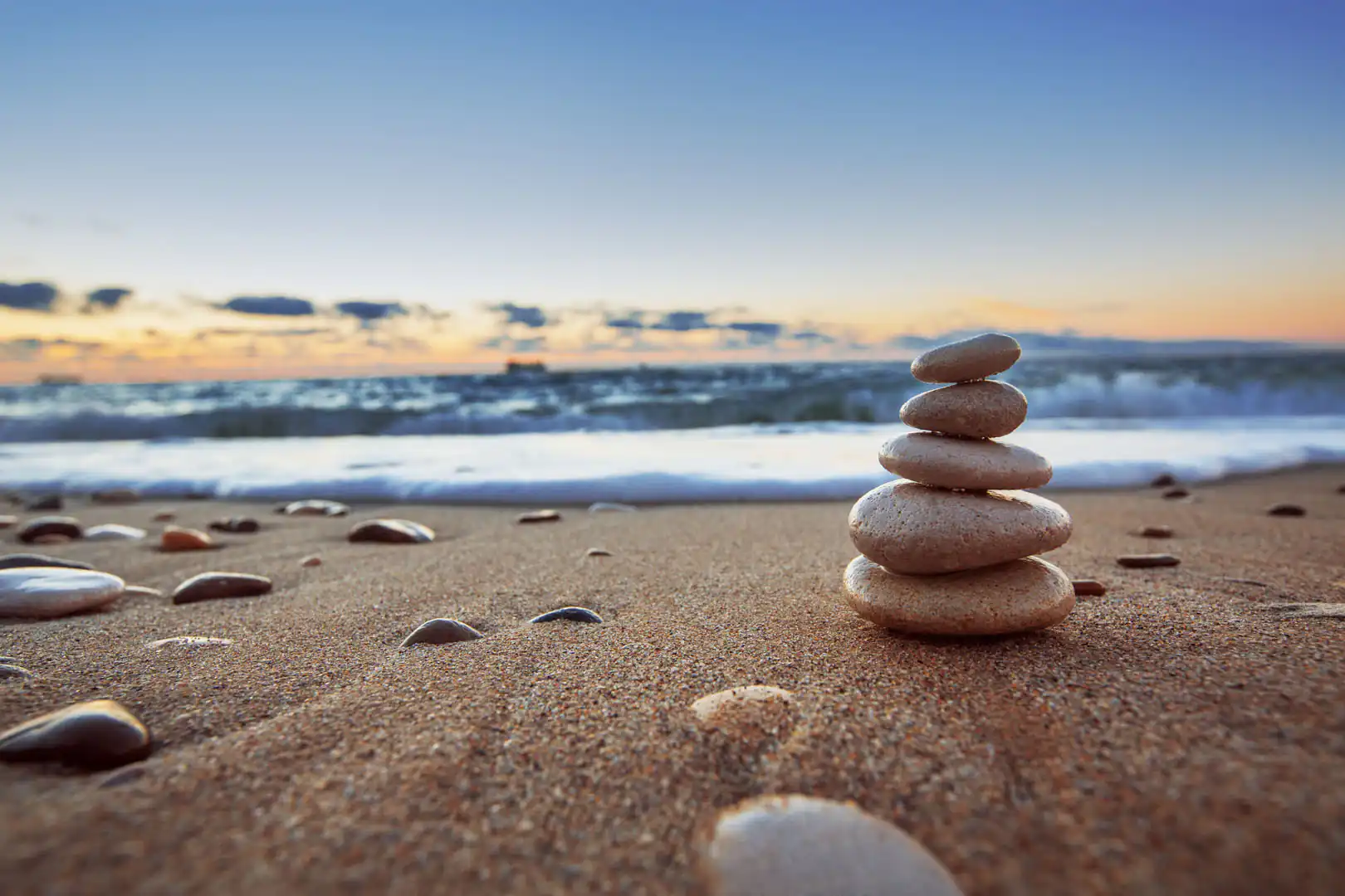 A stack of five smooth stones on a sandy beach in Cornwall at sunset with the sea and sky in the background.